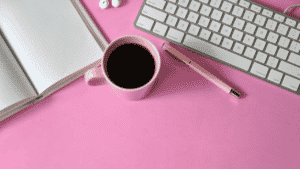 pink desk with coffee, pen, notebook, and keyboard